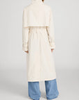A person faces away from the camera wearing FRAME's The Field Trench in light beige with a belted waist and cape detail, paired with wide-leg blue jeans against a plain background.