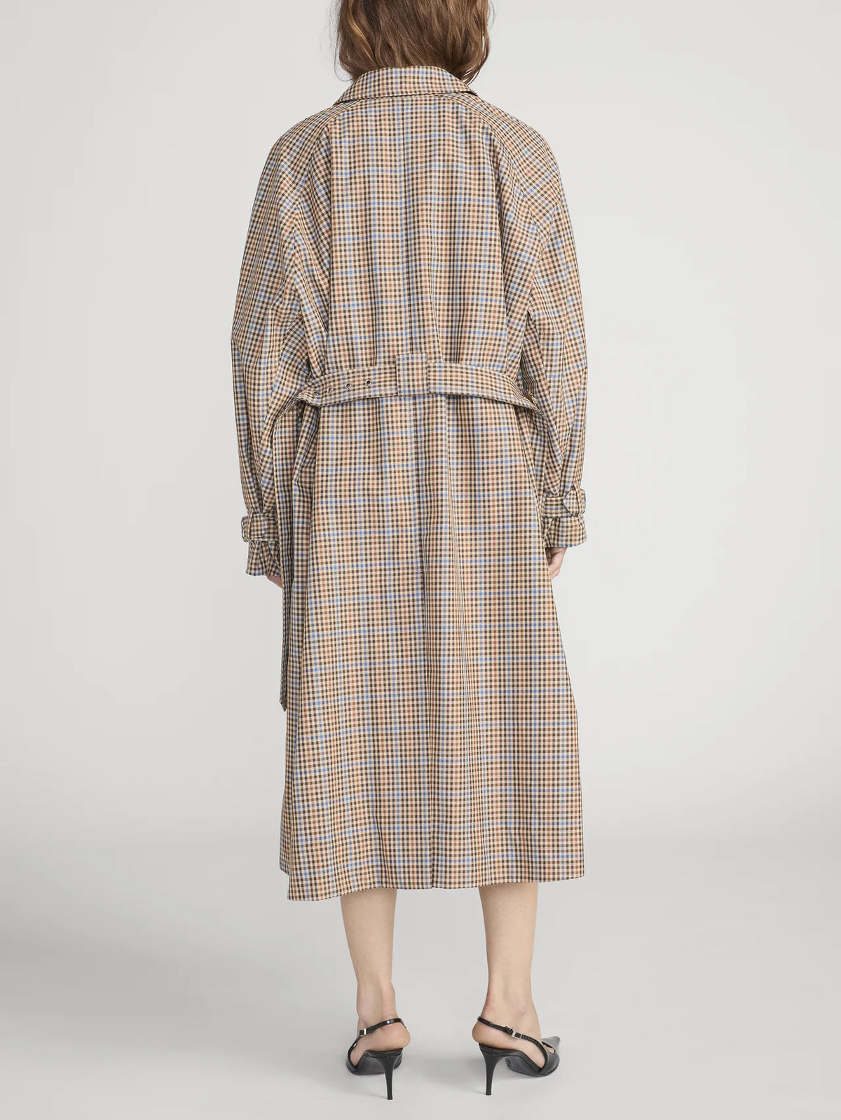 A person wearing The Plaid Mac Coat by FRAME, featuring a tailored finish and belted waist, paired with black heeled sandals, is shown from the back against a plain white background.