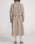 A person wearing The Plaid Mac Coat by FRAME, featuring a tailored finish and belted waist, paired with black heeled sandals, is shown from the back against a plain white background.