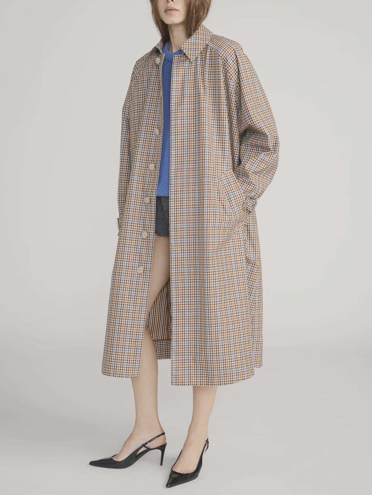 A woman stands against a plain background wearing The Plaid Mac Coat by FRAME over a blue top and dark shorts, paired with black heels. Her hands are in her coat pockets and her face is partially visible.