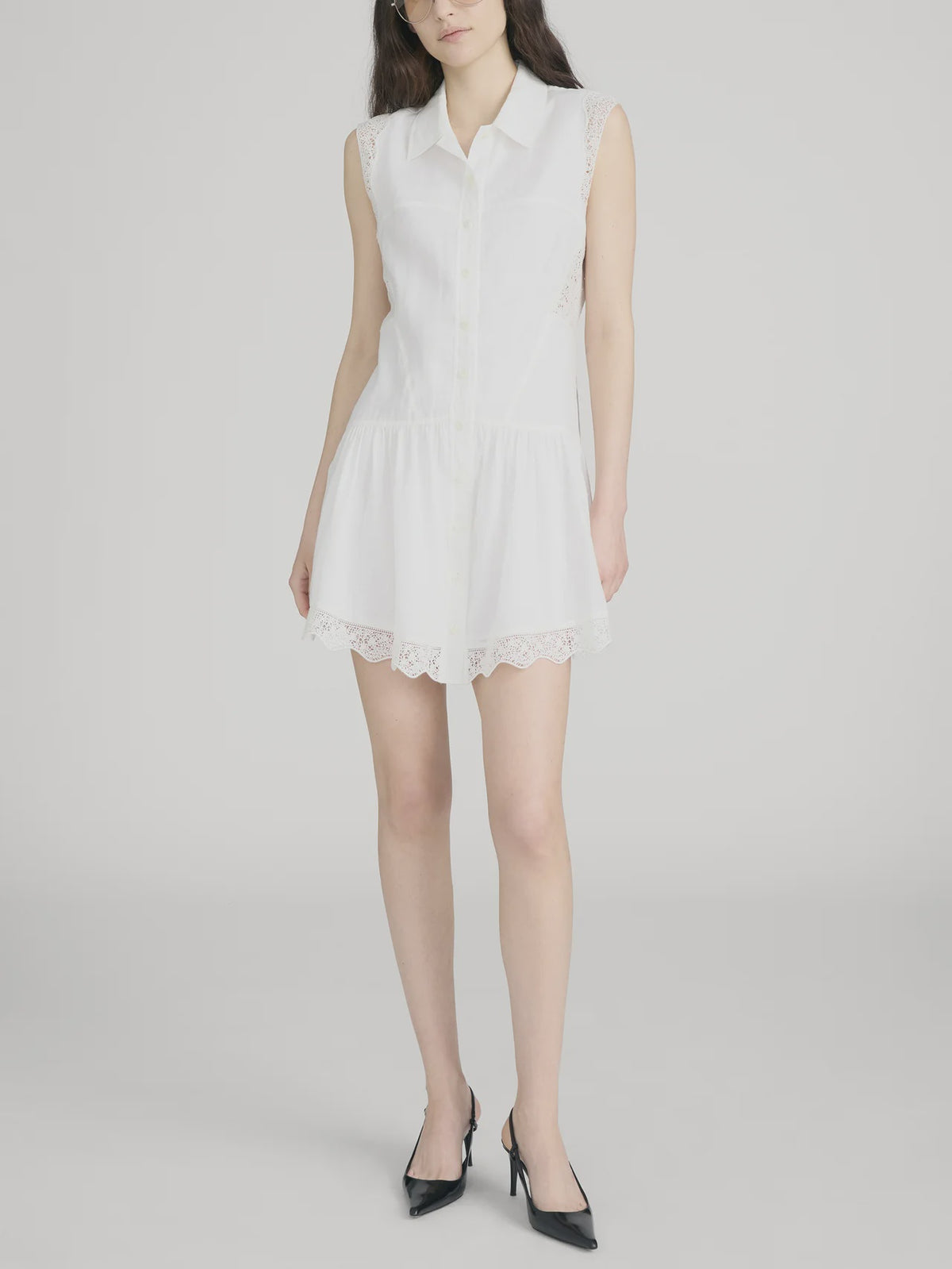 A woman models The Embroidered Day Dress by FRAME—a romantic sleeveless design with a white collar and lace trim at the hem and sides—against a plain light background, styled with black pointed-toe heels.