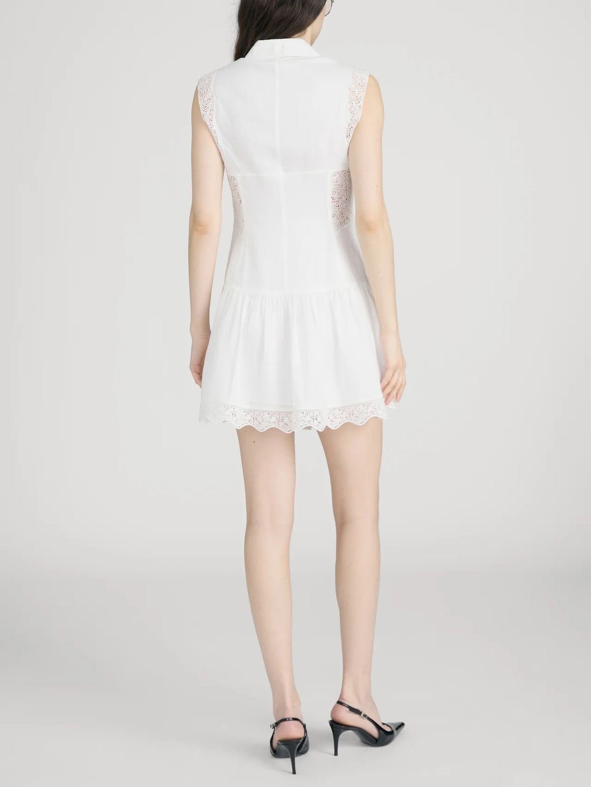 A woman with long dark hair and black heeled sandals stands facing away, wearing FRAME’s Embroidered Day Dress, a romantic sleeveless style featuring lace details and a flared skirt. The background is plain and light-colored.