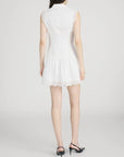 A woman with long dark hair and black heeled sandals stands facing away, wearing FRAME’s Embroidered Day Dress, a romantic sleeveless style featuring lace details and a flared skirt. The background is plain and light-colored.