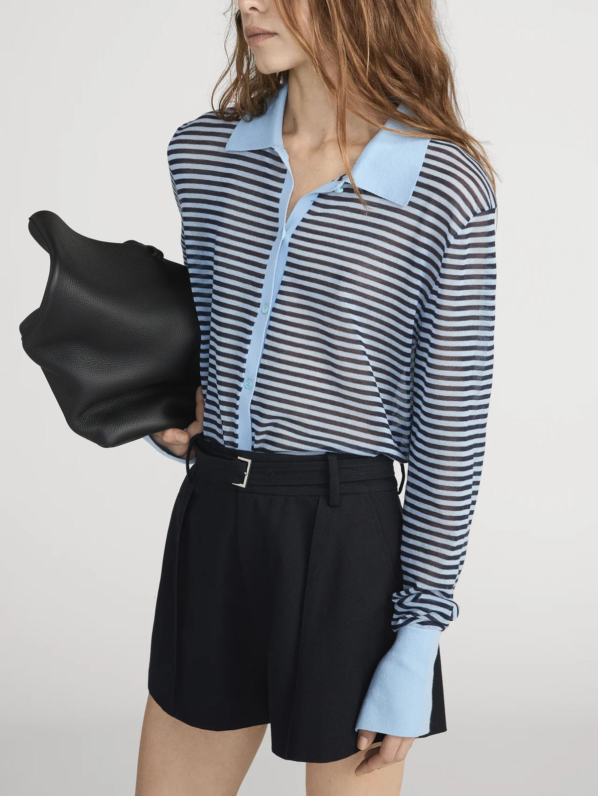 A woman with wavy brown hair wears FRAME’s The Collared Cardi, a sheer blue and navy striped cardigan with a light blue collar and cuffs, paired with high-waisted black shorts and carrying a large black bag.