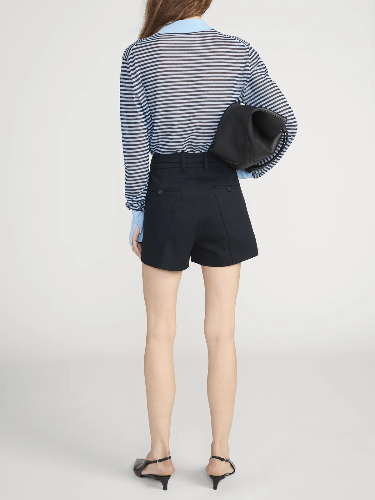 A woman stands with her back to the camera, wearing FRAME's The Collared Cardi in blue and white stripes, paired with high-waisted black shorts, black heeled sandals, and a black clutch under her arm.