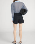 A woman stands with her back to the camera, wearing FRAME's The Collared Cardi in blue and white stripes, paired with high-waisted black shorts, black heeled sandals, and a black clutch under her arm.