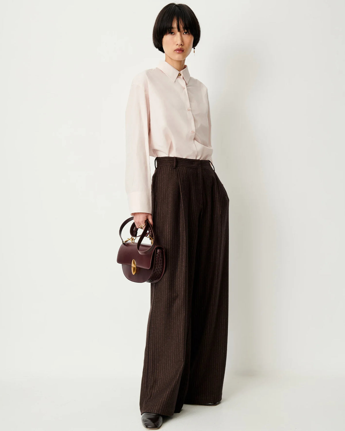 Sessun Orozco Large Pleat Pants styled with a pale pink shirt, shown with a burgundy handbag by a white wall.