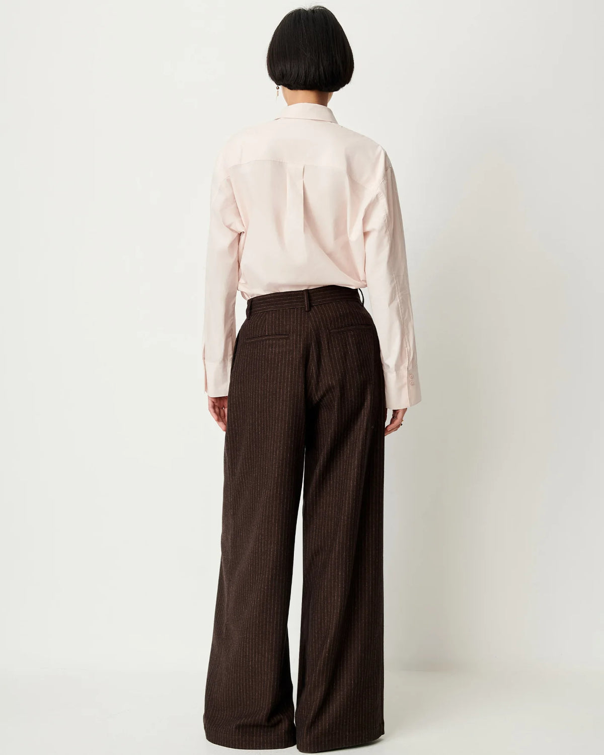 Sessun Orozco pleated large pants and light shirt, model facing away, relaxed modern style.