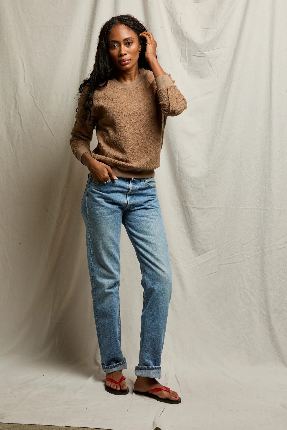 A woman poses before a beige fabric backdrop wearing the Perfectwhitetee Ziggy Reverse LS Sweatshirt in brown, paired with light blue jeans and red sandals. She has long curly hair, looking at the camera with one hand in her pocket and the other touching her hair.