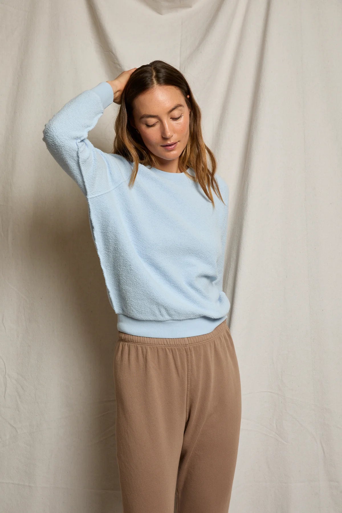 A woman stands before a light beige fabric backdrop, wearing the Perfectwhitetee Ziggy Reverse LS Sweatshirt in light blue and tan pants. She raises one hand to her head and looks down with a relaxed expression.