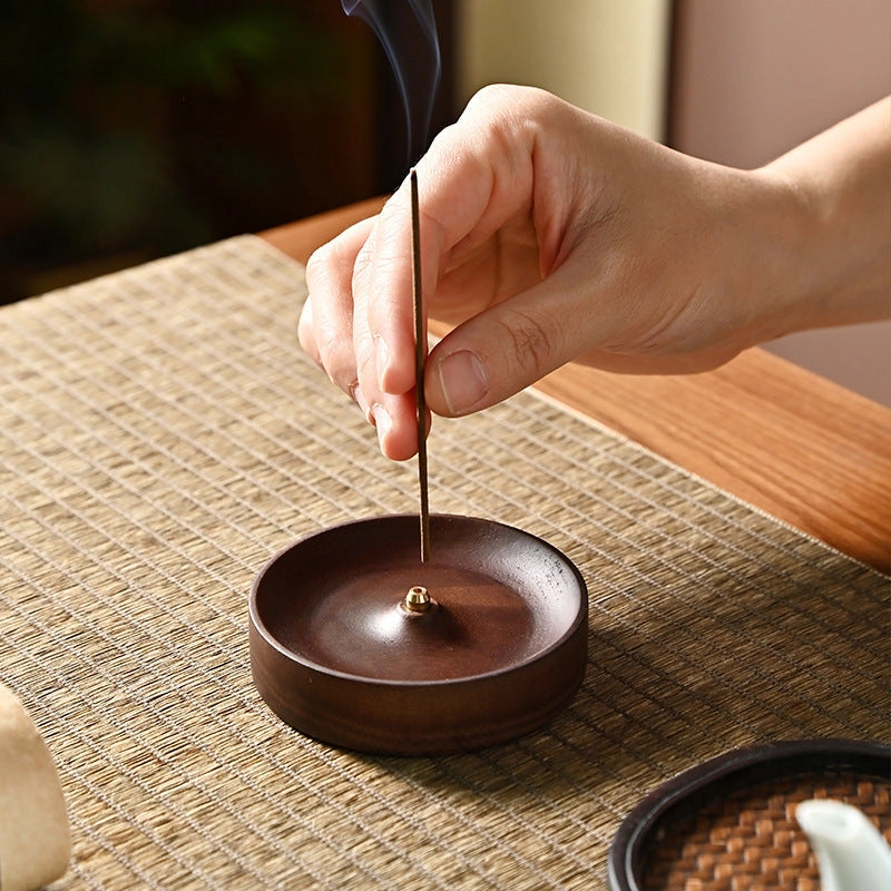 A hand places a lit incense stick into the Faire Walnut Wood Incense Burner on a woven placemat, with smoke rising gently—showcasing traditional Chinese artistry and a unique rustic glaze.