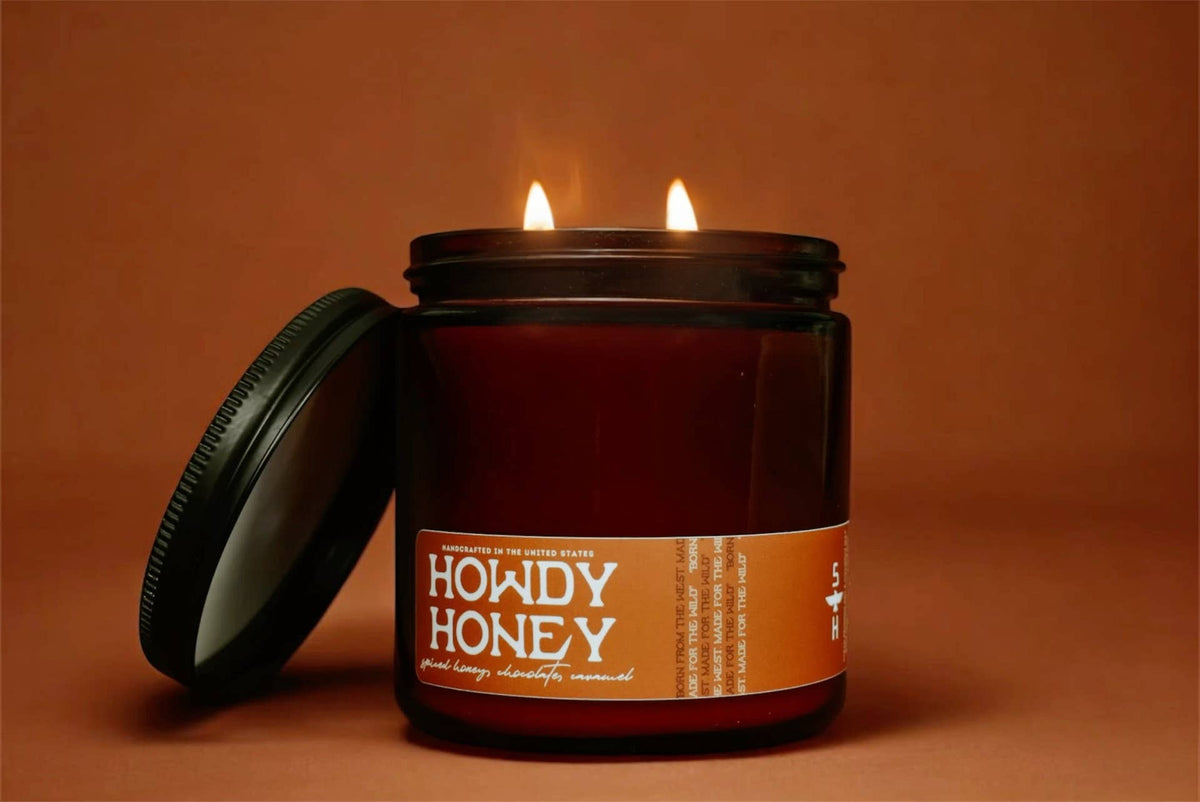 The Faire Howdy Honey Candle is a brown coconut soy wax jar candle with two flames, shown beside its lid on a warm brown background.