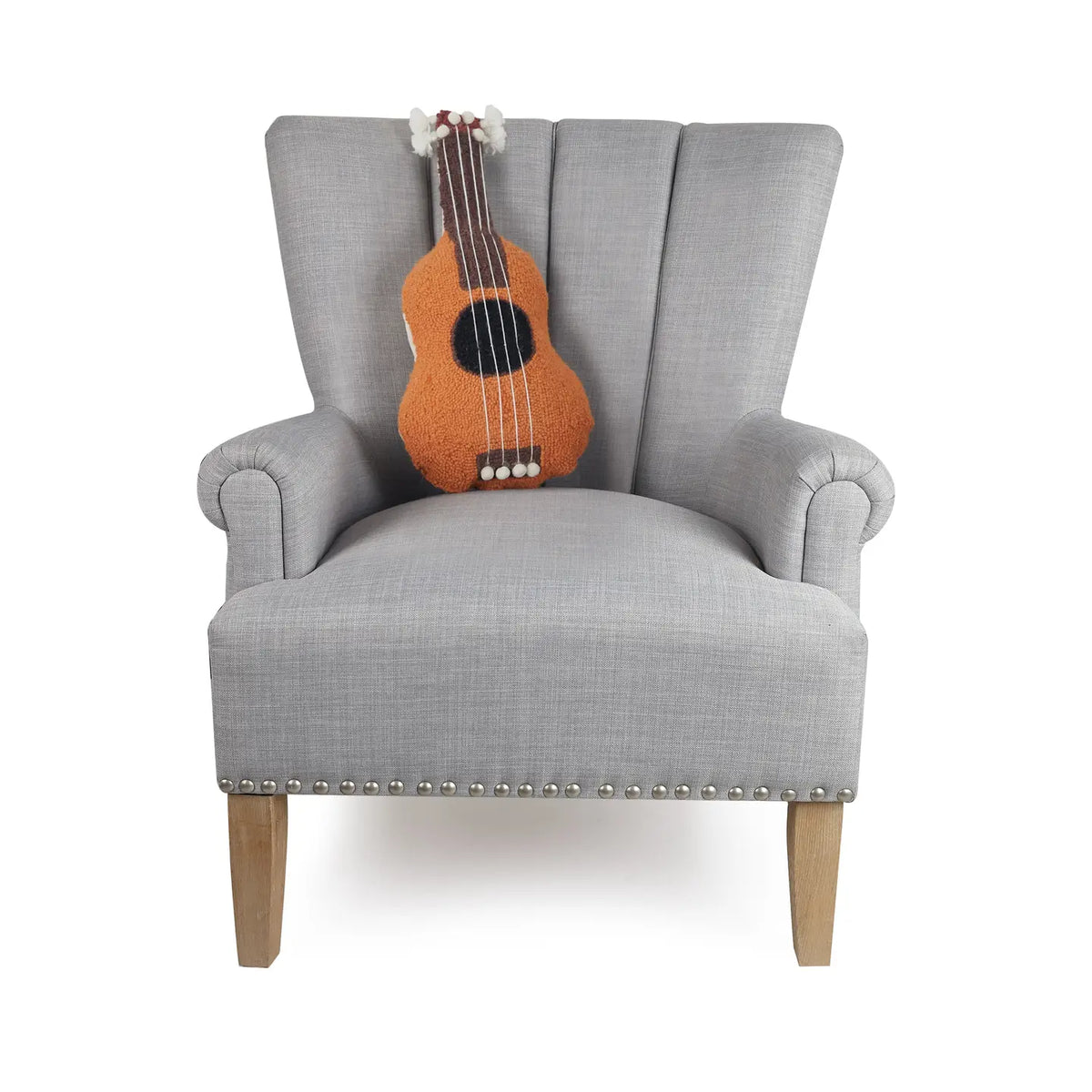 A light gray armchair with wooden legs features the Faire Ukelele Shaped Pillow in plush orange on its backrest and has silver studs along the bottom edge, adding a chic accent ideal for any Jungalow-inspired space.