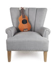 A light gray armchair with wooden legs features the Faire Ukelele Shaped Pillow in plush orange on its backrest and has silver studs along the bottom edge, adding a chic accent ideal for any Jungalow-inspired space.