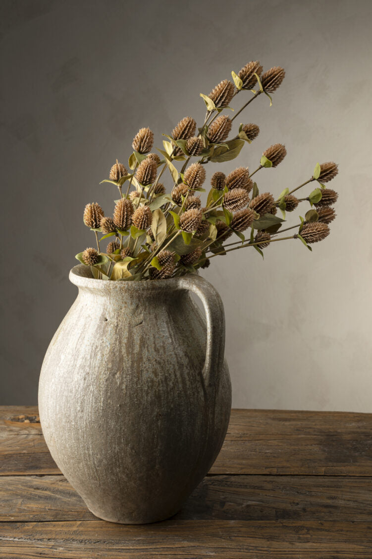 The Indaba Globe Amaranth Spray's sculptural faux botanicals stand in a rustic ceramic vase with a handle, displayed on a weathered wooden table against a neutral, textured background.
