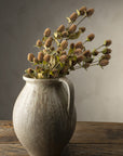 The Indaba Globe Amaranth Spray's sculptural faux botanicals stand in a rustic ceramic vase with a handle, displayed on a weathered wooden table against a neutral, textured background.