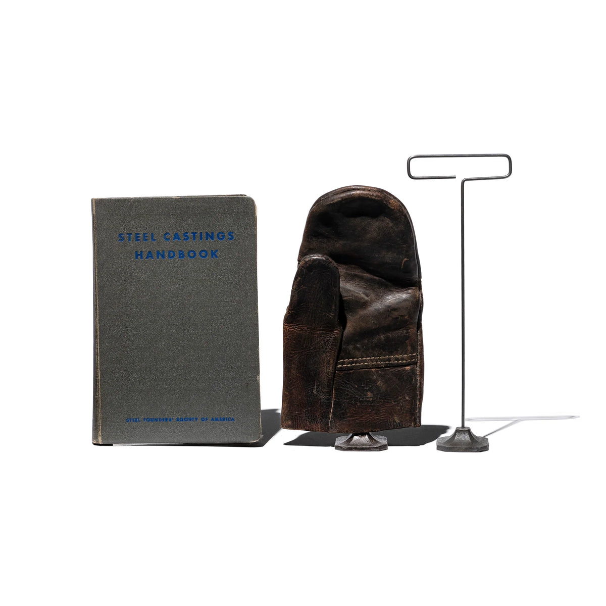 A grey "Steel Castings Handbook," a worn brown leather glove, and a Faire Desk Wire Display Stand L shaped like a 'T' are arranged side by side against a white background.