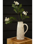 The Indaba Locust Flower Branch features green stems arranged in a rustic white ceramic pitcher with vertical grooves, set on a wooden block against a dark paneled backdrop—an ideal accent decor for any space.