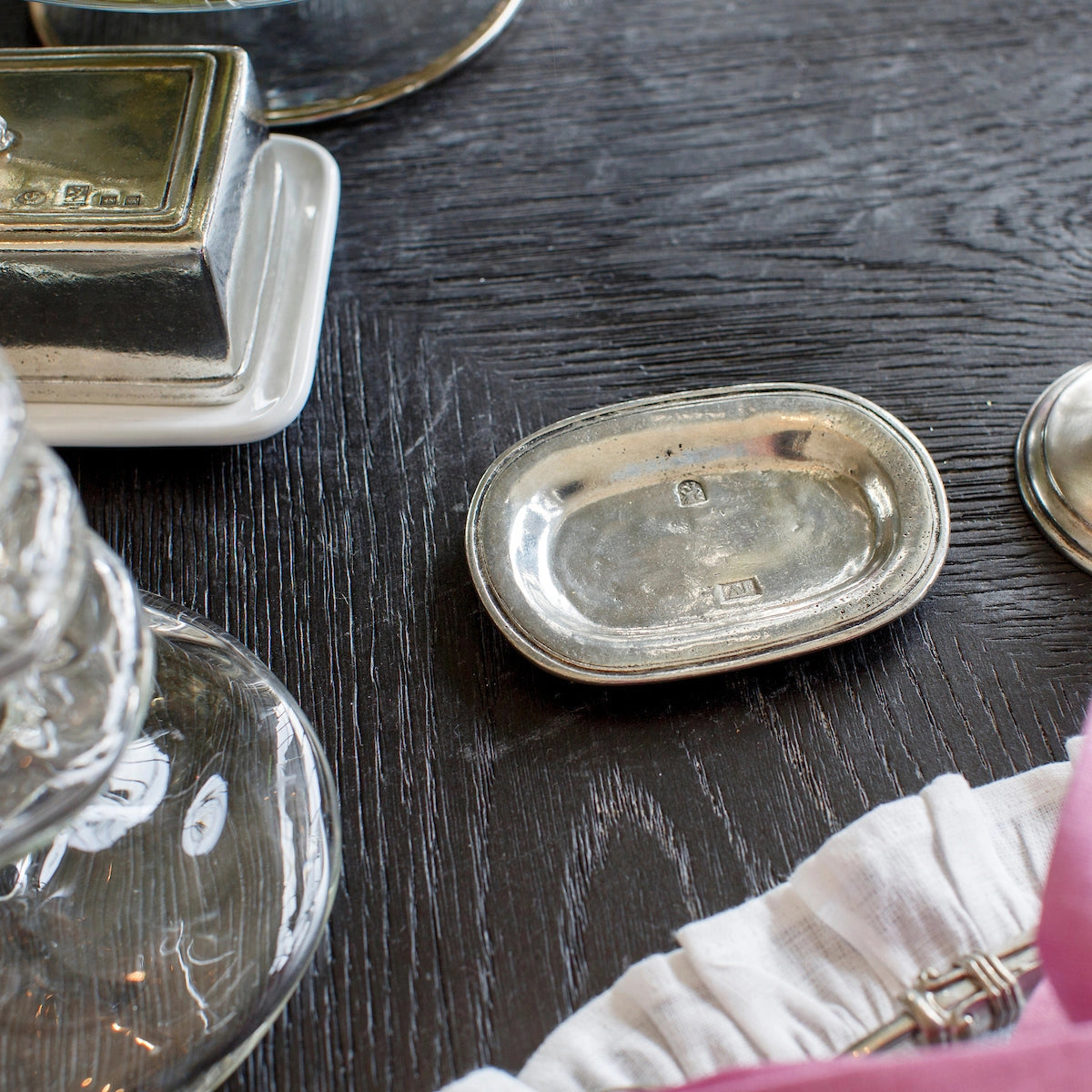 A small, oval Vintage Bedside Tray by Faire, handcrafted from Italian pewter, sits on a dark wooden table beside glassware, a white dish, and a pink and white cloth, reflecting the light.
