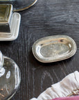 A small, oval Vintage Bedside Tray by Faire, handcrafted from Italian pewter, sits on a dark wooden table beside glassware, a white dish, and a pink and white cloth, reflecting the light.