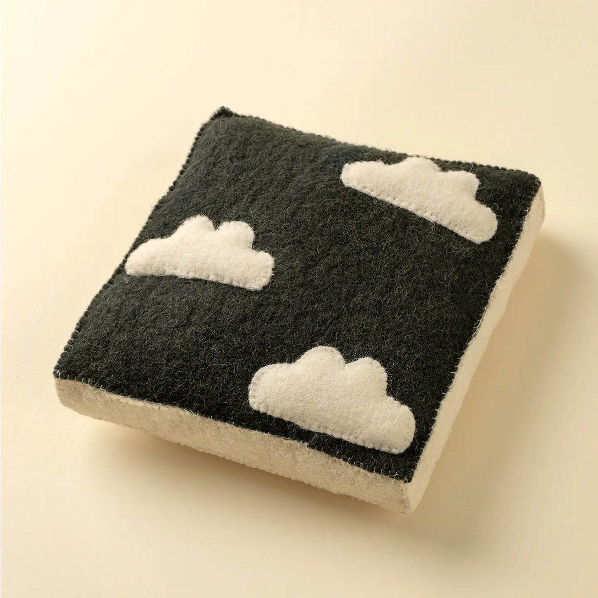 The Indaba Cloudy Day Wall Plaque features three white cloud shapes on a dark square background, ideal as kids' room decor, displayed on a light-colored surface.