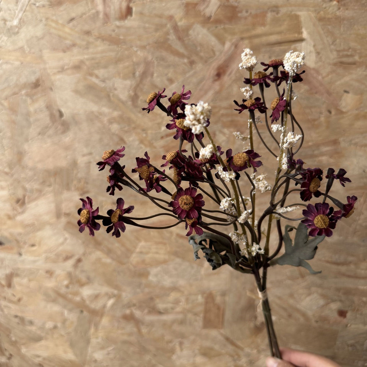 A hand holds the Faire 13'' Faux Daisies Floral Bunch with dark purple and white flowers, including tiny daisies, against a textured brown wooden background.