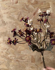 A hand holds the Faire 13'' Faux Daisies Floral Bunch with dark purple and white flowers, including tiny daisies, against a textured brown wooden background.