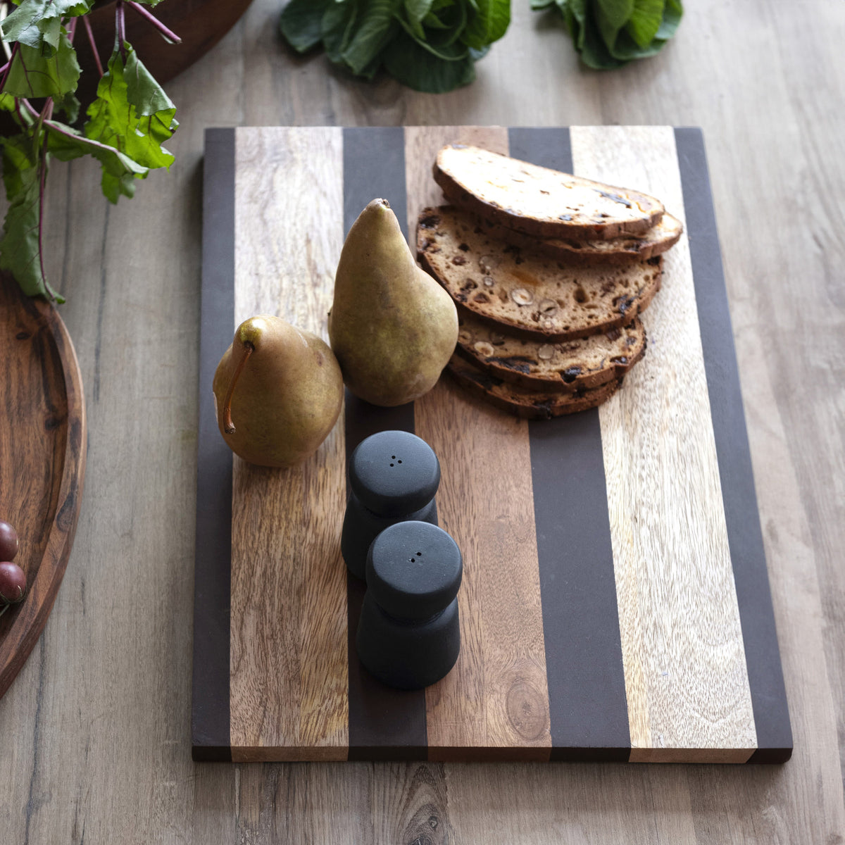 A Bloomingville Mango Wood Serving Board holds two pears, four fruit-studded bread slices, and a black salt and pepper shaker set on a wooden table with leafy greens in the background.