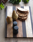 A Bloomingville Mango Wood Serving Board holds two pears, four fruit-studded bread slices, and a black salt and pepper shaker set on a wooden table with leafy greens in the background.