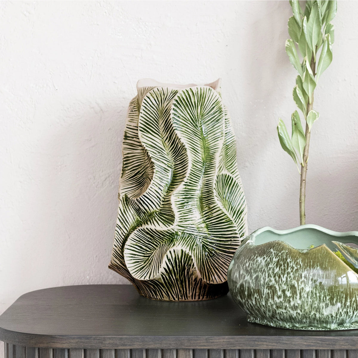 The Bloomingville Green Floral Texture Vase (round stone vase) with a green palm leaf pattern sits on a gray console table beside a green glazed bowl and a tall leafy stem, set against a textured white wall.