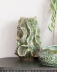 The Bloomingville Green Floral Texture Vase (round stone vase) with a green palm leaf pattern sits on a gray console table beside a green glazed bowl and a tall leafy stem, set against a textured white wall.