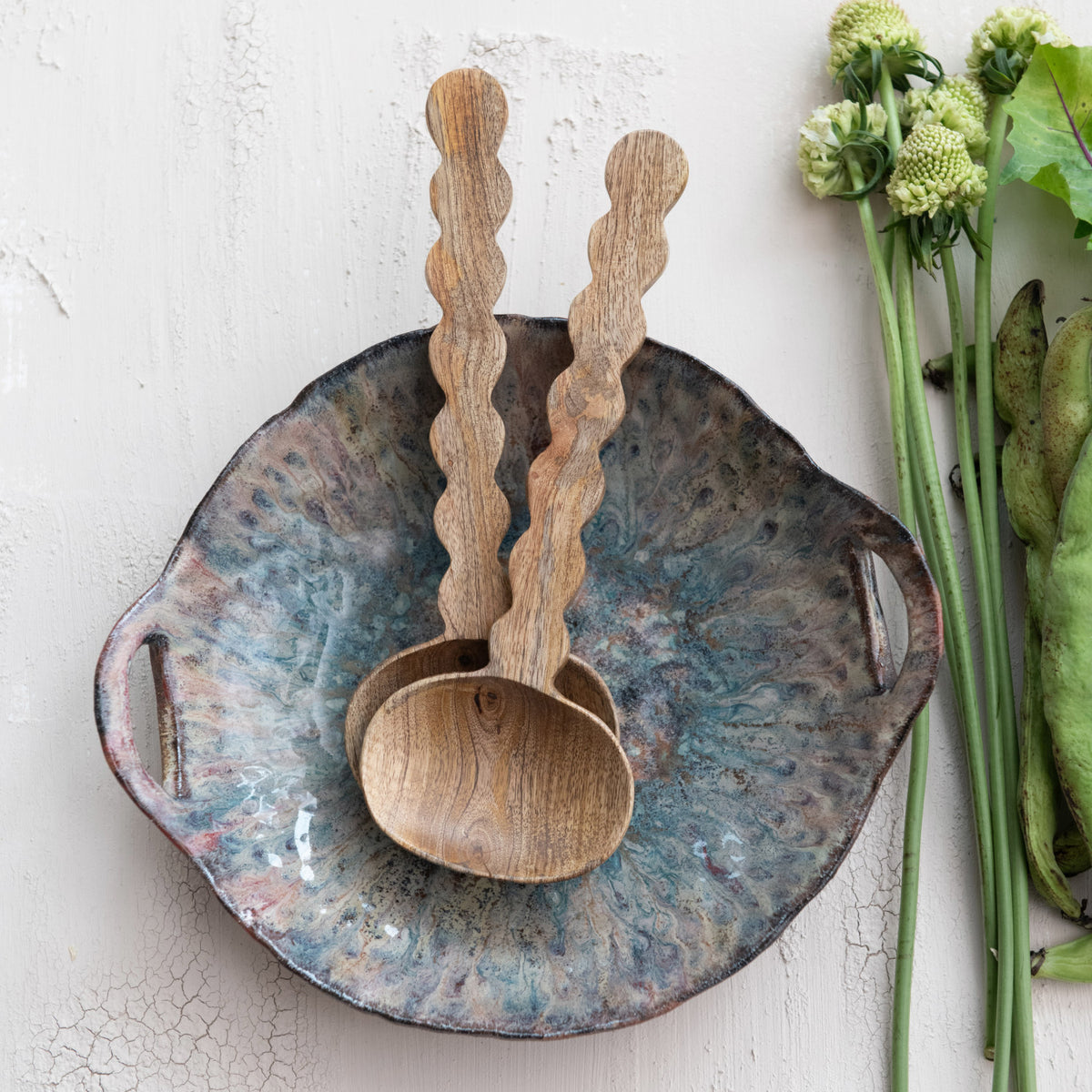 Bloomingville Mango Salad Servers with scalloped handles are paired with a textured ceramic bowl, displayed on a light surface alongside green vegetables and white flower buds.