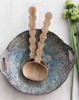 Bloomingville Mango Salad Servers with scalloped handles are paired with a textured ceramic bowl, displayed on a light surface alongside green vegetables and white flower buds.