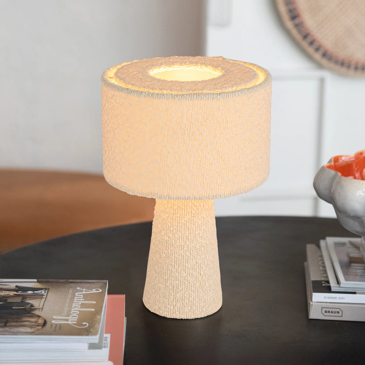 The Bouclé Ivory Fabric Table Lamp by Bloomingville, featuring a textured base and cylindrical shade, glows warmly on a dark round table beside stacked magazines and a ceramic bowl in a cozy indoor space.