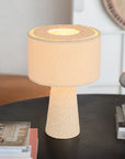 The Bouclé Ivory Fabric Table Lamp by Bloomingville, featuring a textured base and cylindrical shade, glows warmly on a dark round table beside stacked magazines and a ceramic bowl in a cozy indoor space.