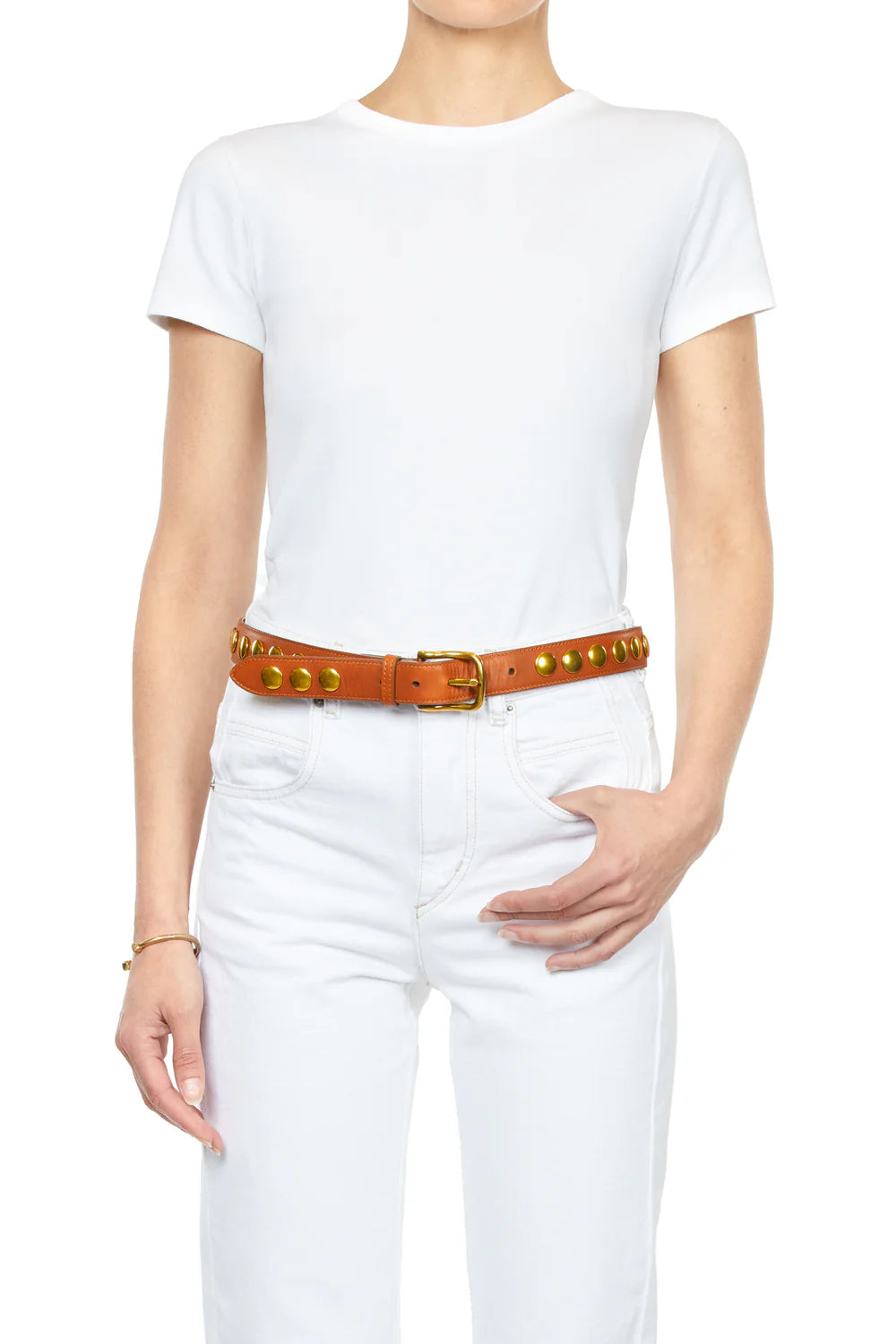 Dressed in a white t-shirt and jeans, a person poses with one hand on their waist, flaunting Jerome Dreyfuss's Festival Ceinture — a chic tan cowskin leather belt embellished with gold studs and a shiny gold buckle, while their other arm hangs relaxed by their side.