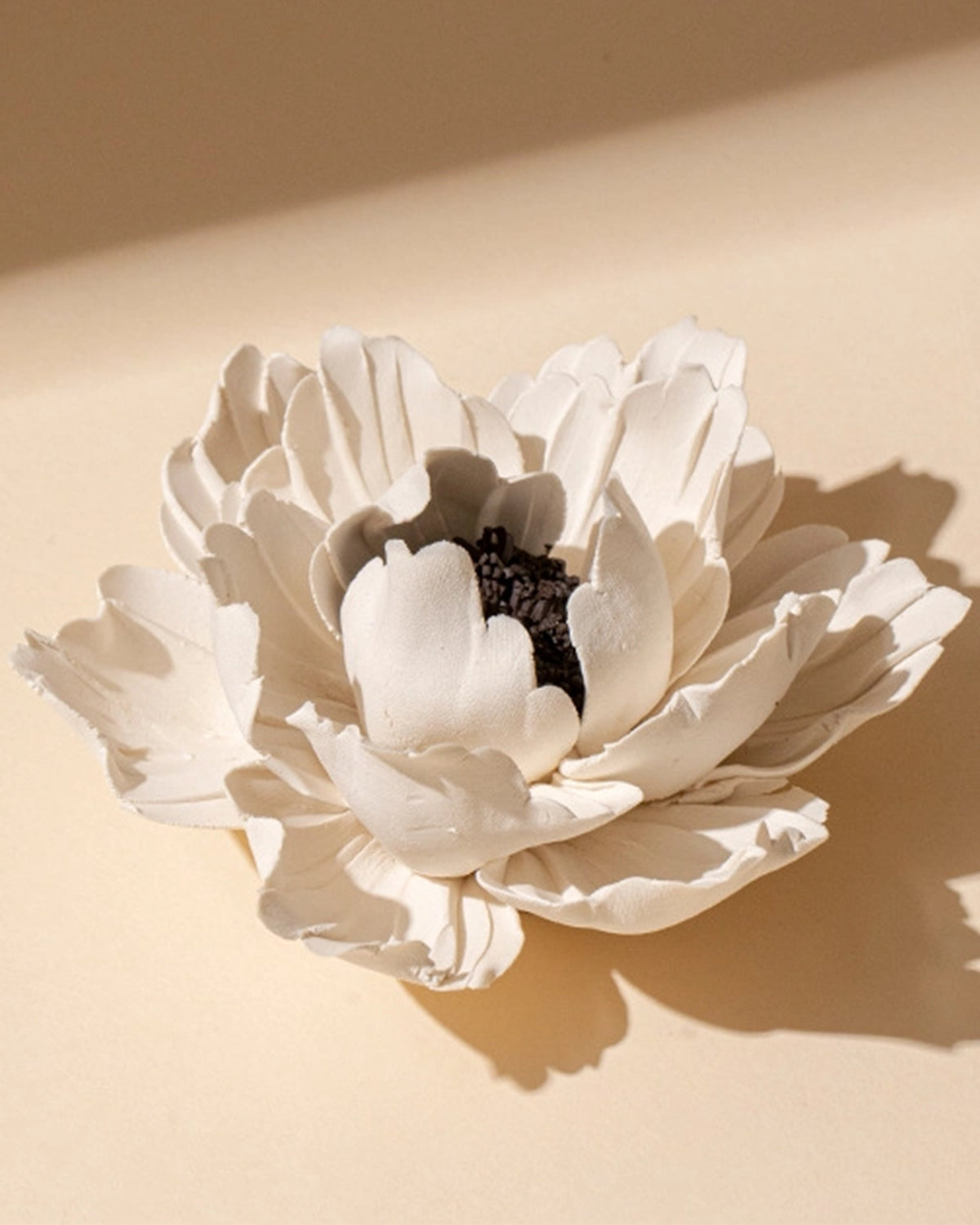The Faire Ceramic Anemone Incense Holder features delicate, layered white petals and a dark textured center, evoking traditional Chinese artistry. It rests on a soft beige surface under gentle lighting.