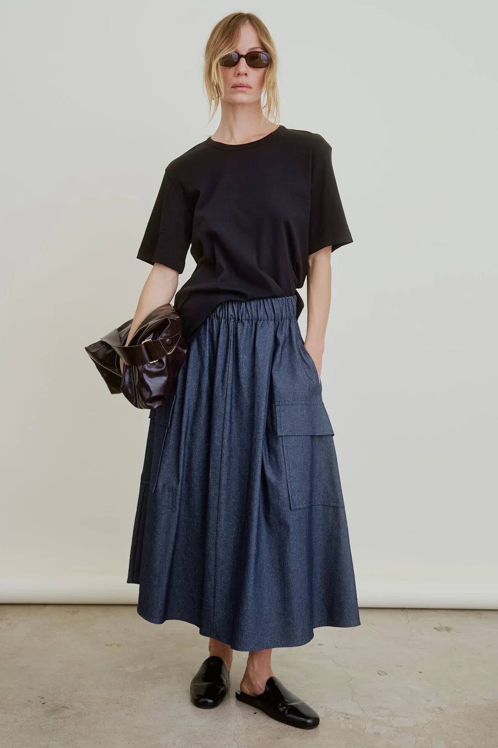 Wearing sunglasses, a loose black t-shirt, Aquarius Cocktail’s Le Cargo Skirt in blue Japanese cotton poplin with oversized pockets, and black slip-on shoes, this person stands against a plain background holding a shiny black bag.