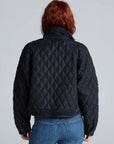 A person with red hair, seen from behind, wears the ASKK Bleecker Street Liner— a black, quilted, cropped jacket—paired with blue jeans against a plain light gray background.