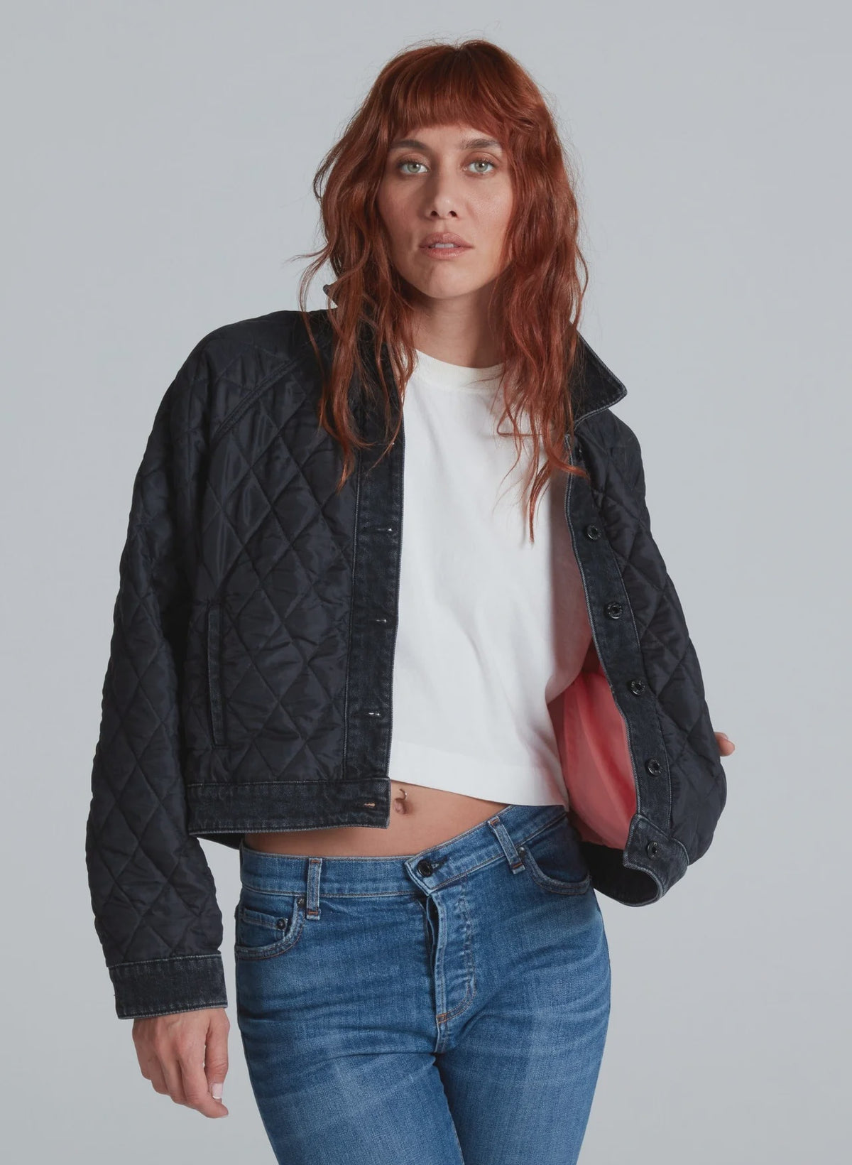 A person with long, wavy red hair wears the ASKK Bleecker Street Liner—a black quilted, cropped jacket with one side open to show its pink lining—paired with a white crop top and blue jeans against a plain light gray background.