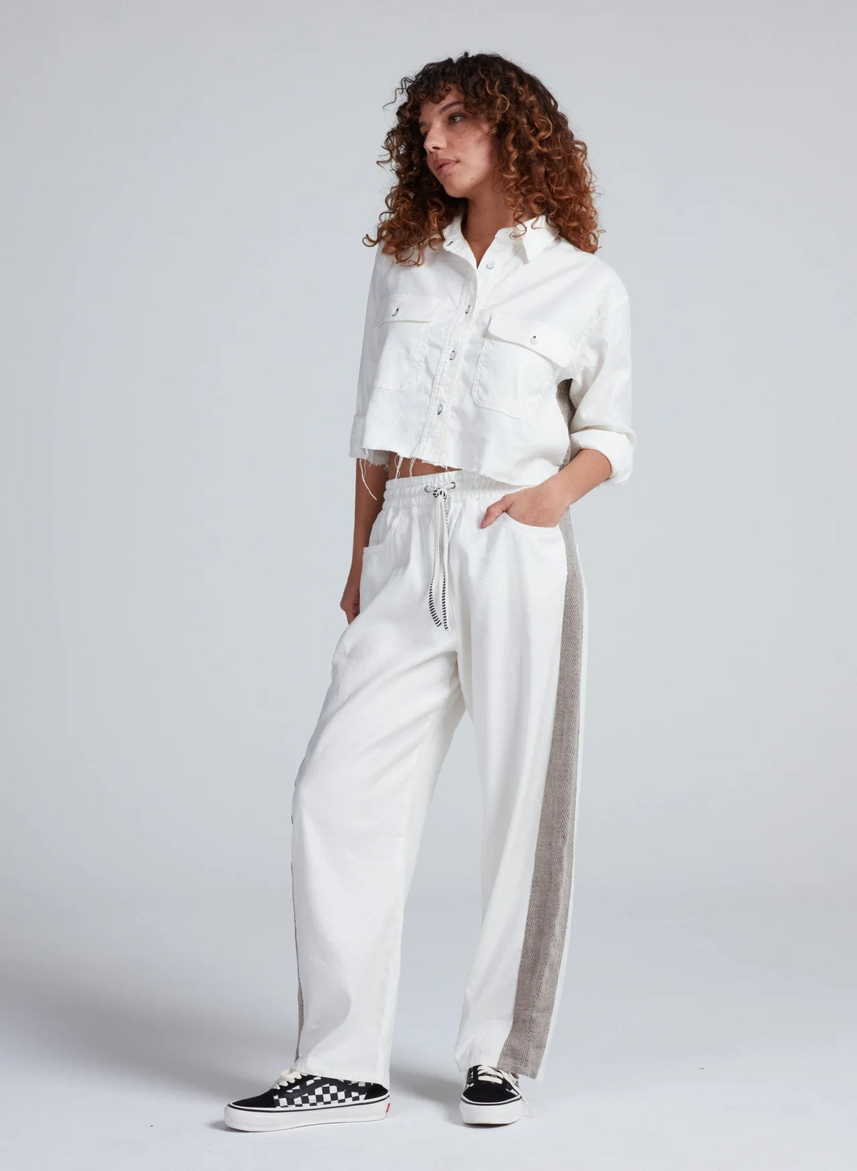 A woman with curly hair wears the ASKK Cancun Pant—white wide-leg drawstring pants with a gray side stripe and elastic waistband—and a cropped white button-up jacket, standing with one hand in her pocket against a plain background.