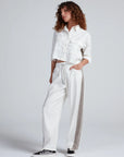 A woman with curly hair wears the ASKK Cancun Pant—white wide-leg drawstring pants with a gray side stripe and elastic waistband—and a cropped white button-up jacket, standing with one hand in her pocket against a plain background.