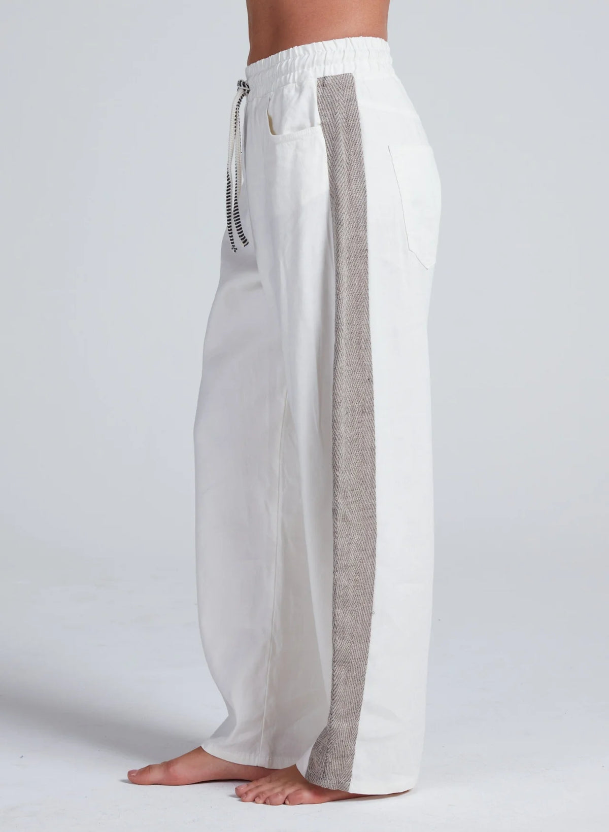 A barefoot model is shown from the side against a plain light background wearing the ASKK Cancun Pant: loose-fitting, wide leg white pants with a gray side stripe, elastic waistband, and side pockets.