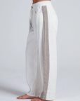 A barefoot model is shown from the side against a plain light background wearing the ASKK Cancun Pant: loose-fitting, wide leg white pants with a gray side stripe, elastic waistband, and side pockets.