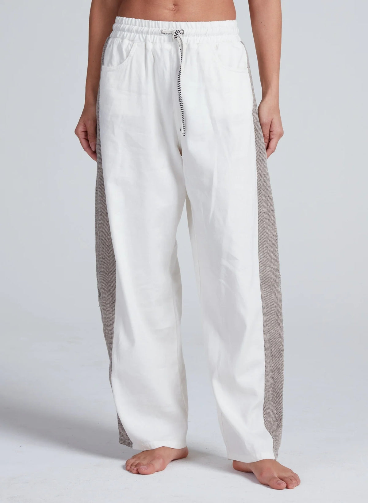 A person stands barefoot in ASKK's Cancun Pant, featuring a wide-leg silhouette, white fabric with gray vertical side panels, and an elastic drawstring waistband, photographed against a plain light background.