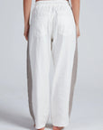 A person stands barefoot with their back to the camera, wearing ASKK’s Cancun Pant—loose white wide leg pants with gray textured side panels, large back pockets, and an elastic waistband—set against a plain light background.