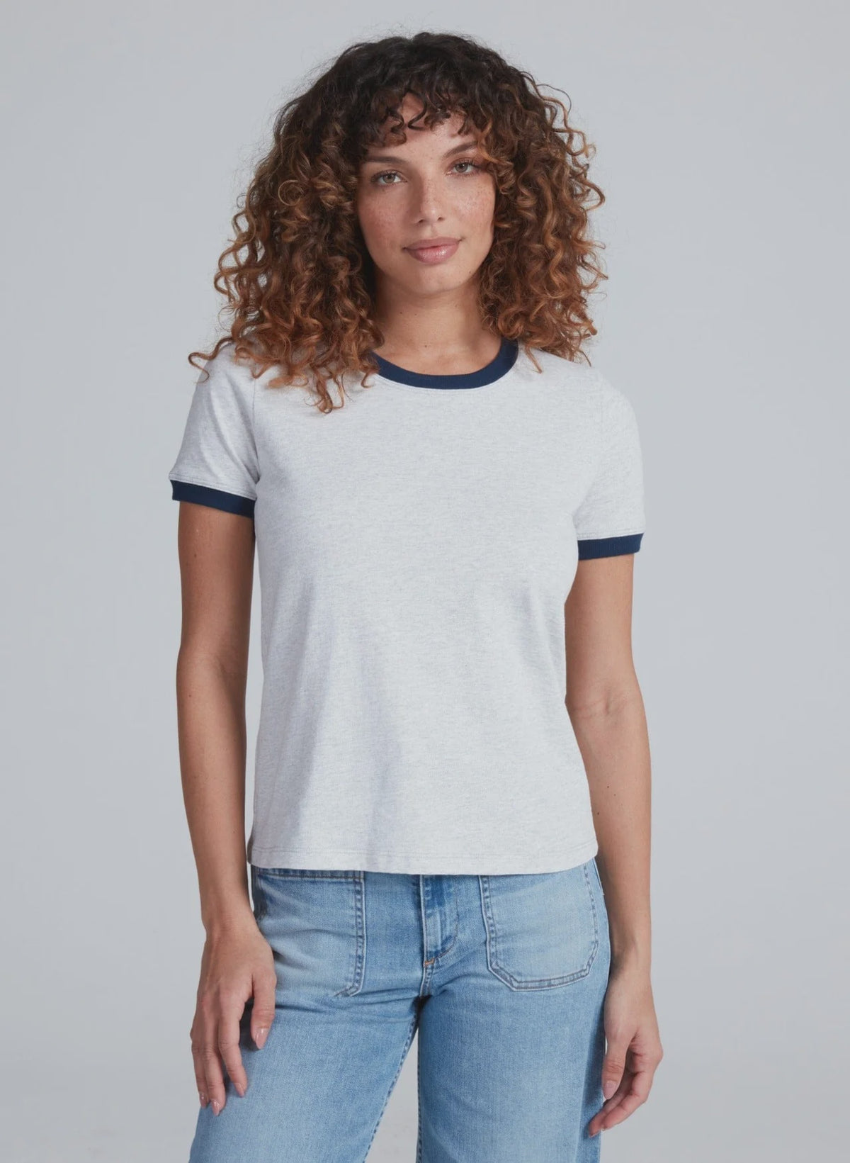 A woman with curly brown hair wears the ASKK Classic Ringer Tee, made from American knit cotton, paired with blue jeans, standing facing forward against a plain light gray background.