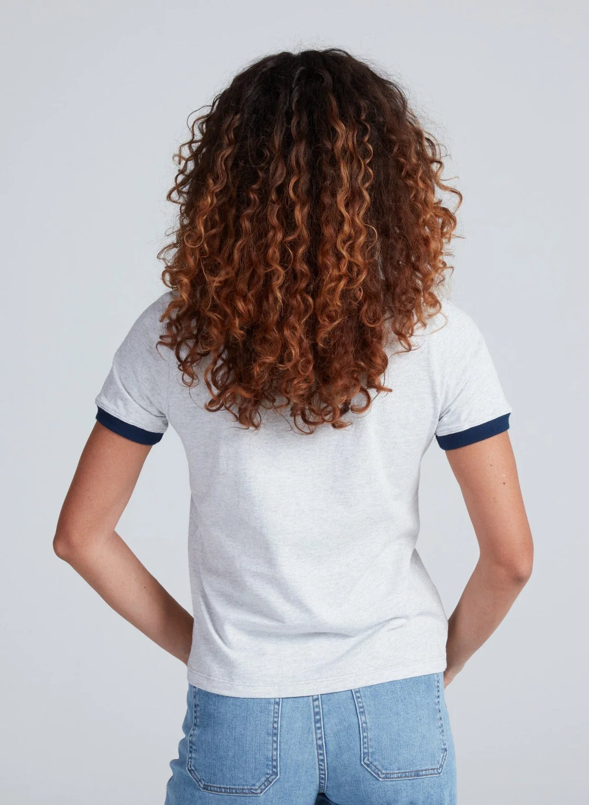 Seen from behind, a person with curly, shoulder-length brown hair wears ASKK's Classic Ringer Tee and blue jeans, standing against a simple light background.