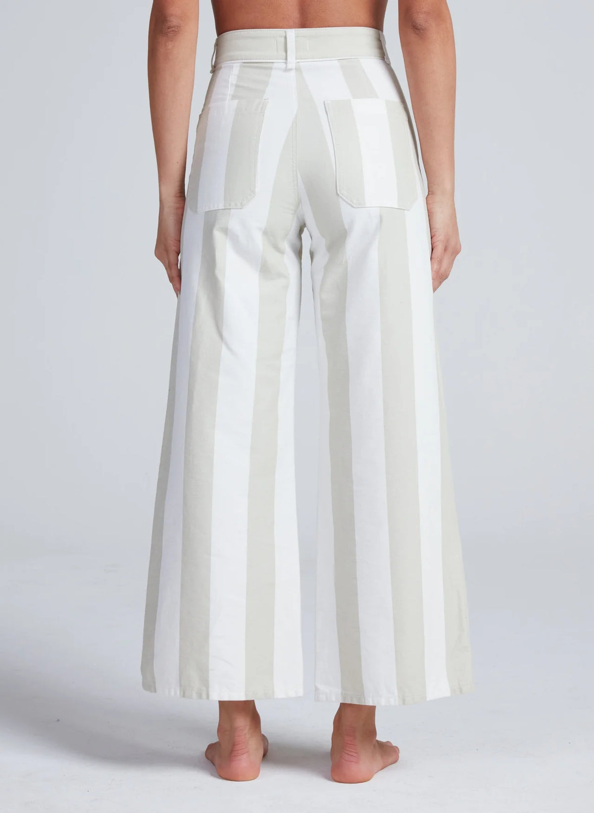 A person stands barefoot, facing away, wearing ASKK's Cropped Sailor high-rise stretch twill wide-leg pants with vertical white and light beige stripes and large back pockets. The background is plain light gray.