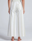 A person stands barefoot, facing away, wearing ASKK's Cropped Sailor high-rise stretch twill wide-leg pants with vertical white and light beige stripes and large back pockets. The background is plain light gray.